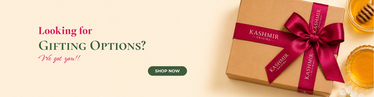 A promotional website banner on a light beige background. On the left, the text reads 'Looking for Gifting Options? We got you!!' above a dark green 'Shop Now' button. On the right is a brown kraft gift box tied with a magenta ribbon printed with the words 'Kashmir Origins', positioned next to two small glass bowls of honey, a wooden honey dipper, and a small white flower.