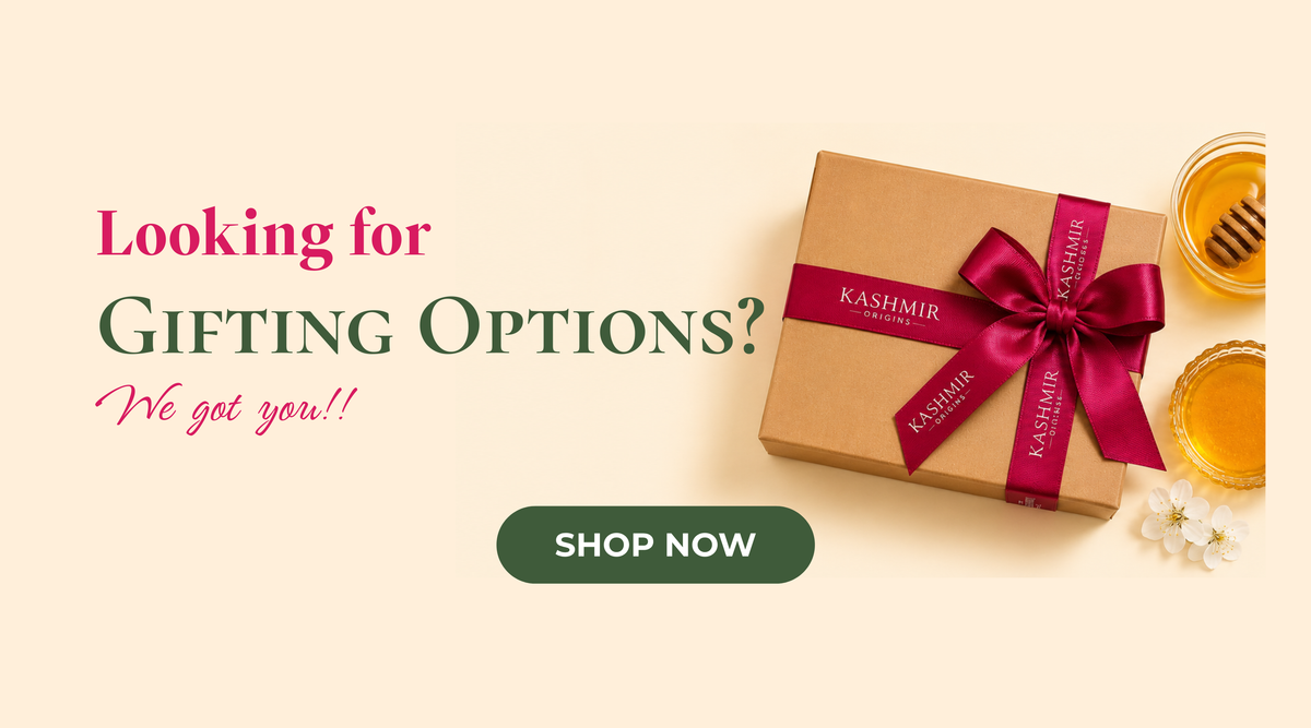 A promotional website banner on a light beige background. On the left, the text reads 'Looking for Gifting Options? We got you!!' above a dark green 'Shop Now' button. On the right is a brown kraft gift box tied with a magenta ribbon printed with the words 'Kashmir Origins', positioned next to two small glass bowls of honey, a wooden honey dipper, and a small white flower